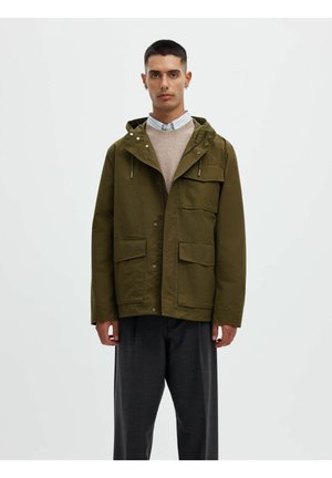 Homme portant une veste à capuche vert olive par-dessus un pull beige et une chemise col, debout devant un fond clair uni.