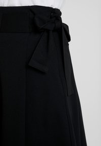 Pantalones negros de cintura alta con un cinturón de tela atado en un lazo, con textura suave y cierre de cremallera lateral.