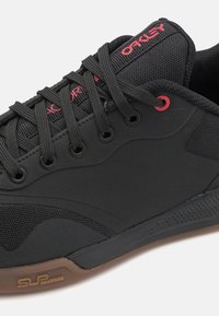 Black athletic shoe featuring a textured surface, lace-up design, reinforced eyelets, contrasting red accents, and a gum rubber outsole.