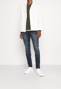 Chaqueta blanca de botón sobre un suéter verde oscuro. Jeans oscuros de corte slim y zapatillas blancas completan el atuendo. Diseño limpio y moderno.