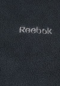 Svart texturerad tyg med en vit broderad "Reebok"-logotyp centrerad på materialet.