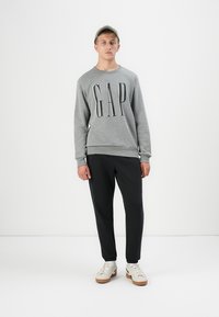 Sudadera gris con la palabra "GAP" impresa en grandes letras negras, combinada con joggers negros y zapatillas blancas. El modelo también lleva puesta una gorra gris.