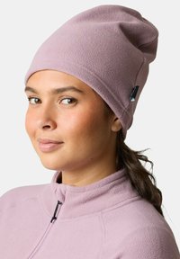 Vaaleanpunainen fleece-pipo, jossa on rento muotoilu ja pieni musta logotagi sivussa. Nainen käyttää sitä lähikuvassa.