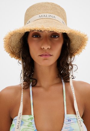 Νεαρή γυναίκα με σγουρά μαλλιά φοράει ψάθινο καπέλο με κορδέλα «MALINA» και παστέλ λουλουδάτο τοπ με λαιμόκοψη halter, κοιτάζοντας μπροστά σε λευκό φόντο.