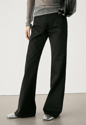 Persona che indossa pantaloni neri a gamba larga, top grigio a maniche lunghe, scarpe argento con punta quadrata e tiene una borsa nera di lato.