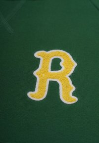 Grön tygskjorta med ett stort, fluffigt gult broderat "R" som är omgärdat av vitt. Slät textur med rundad halsringning.