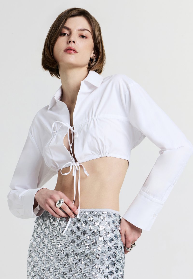 Camicia bianca corta con maniche lunghe, dettaglio annodato davanti, texture liscia. Abbinata a una gonna argentata con paillettes, che mette in evidenza i riflessi.