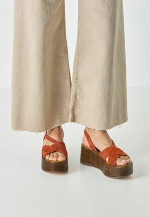 Sandali con platform marroni con suola testurizzata e design a incrocio in suede rosso. Indossati con pantaloni beige a gamba larga con orlo sfrangiato.