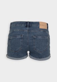 Denim-shortsit tumman sinisen pesun kanssa, joissa on tekonahkainen tagi, käännetyt lahkeet ja kaksi takataskua. Ompeleet ovat näkyvissä vyötärön ympärillä.