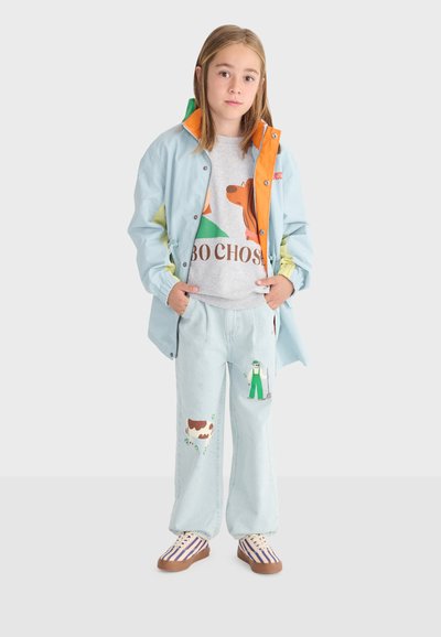 Pige med glat hår iført lyseblå jakke med orange foring, grå sweatshirt med hundegrafik, lyse jeans med bonde- og ko-tryk, stribede sko.