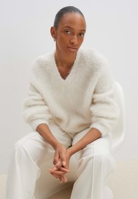 Weißer, flauschiger Pullover mit V-Ausschnitt und übergroßen Ärmeln, kombiniert mit cremefarbenen Hosen, die eine glatte Textur und einen lässigen Schnitt aufweisen.