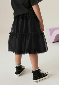 Jupe en tulle noir avec des volants superposés, taille élastique, portée avec des baskets montantes noires et des chaussettes grises. Fond uni.