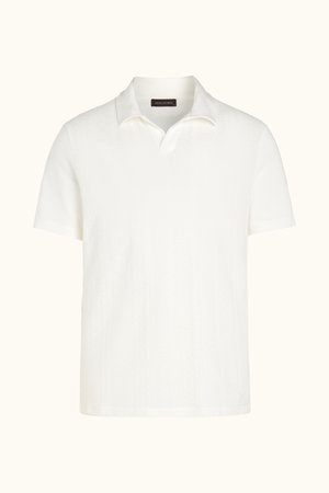 Uomo in piedi con una mano in tasca, indossa una polo bianca a maniche corte in maglia, pantaloncini neri e mocassini neri su sfondo semplice.
