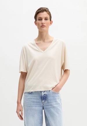 Femme portant un t-shirt beige à manches courtes et col en V ample, ainsi qu'un jean bleu clair, sur un fond blanc uni.