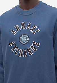 Άνδρας με μπλε φούτερ με λευκό και μαύρο κείμενο "Armani Exchange" και κυκλικό έμβλημα Μιλάνου στο στήθος.