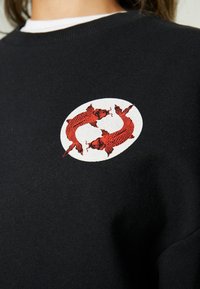 Svart sweatshirts med en röd broderad design av två koi-fiskar inom en vit oval, mot en texturerad tygbakgrund.