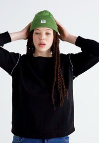 Black sweatshirt with decorative details along sleeves, paired with a green knit beanie featuring a patch. Model has long, curly hair.