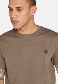 T-shirt in cotone marrone con maniche corte e scollatura a girocollo. Presenta un piccolo logo di un albero ricamato sul lato sinistro del petto.