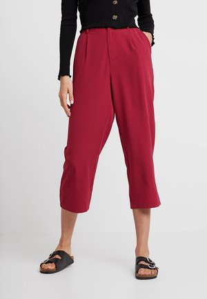 Pantalons bourgogne raccourcis avec une taille haute, des plis et une texture lisse. Portés avec des sandales noires dotées d'une grande boucle en argent.