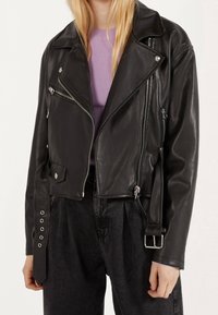 Vrouw in een zwart leren motorjack met zilveren ritsen, een riem, en een lavendelgekleurde geribde trui eronder.