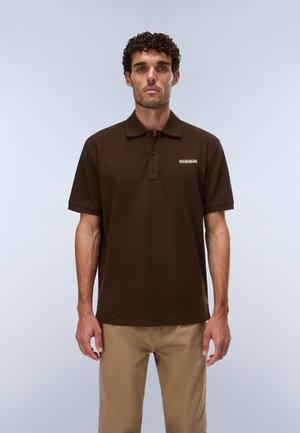 Uomo con capelli corti e ricci, che indossa una polo marrone scuro e pantaloni beige, in piedi davanti a uno sfondo grigio chiaro.
