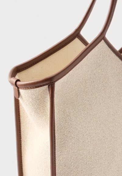 Sac fourre-tout en tissu beige texturé avec garniture et poignées en cuir brun lisse, présenté en gros plan sur un fond clair.