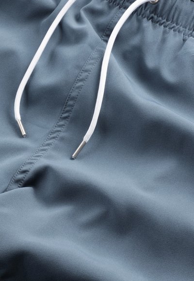 Shorts de bain bleus en tissu lisse avec un cordon de serrage, présentant des cordons blancs et des embouts métalliques, mettant en avant une texture douce.