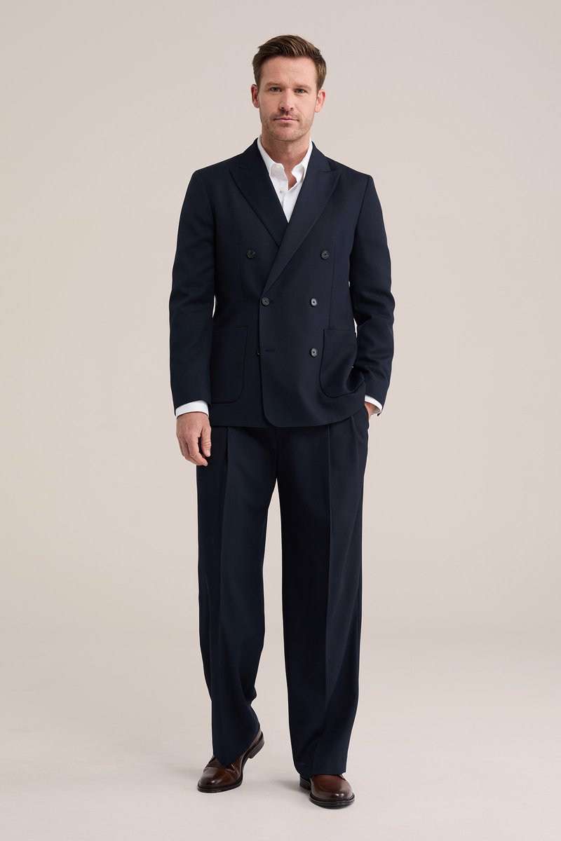 Hombre de pie vistiendo un traje cruzado azul marino con pantalones de pierna ancha, camisa blanca y zapatos marrones sobre un fondo beige liso.