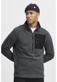 Pullover in felpa grigia con una tasca sul petto nera con zip, colletto a mezza zip e tasche laterali. Texture morbida, taglio dritto e polsini a coste.