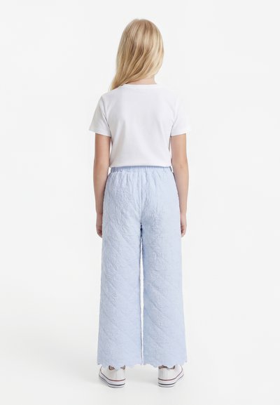 Svetlovlasé dieťa stojí s chrbtom, má na sebe bielu košeľu s krátkym rukávom, svetlomodré široké textúrované nohavice a biele tenisky.