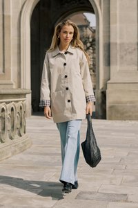 Jeune femme marchant en plein air près d'une arche en pierre, portant un manteau beige, un jean bleu clair, des bottes noires, et tenant un grand sac noir tressé.