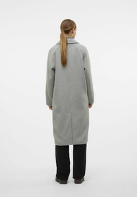 Cappotto grigio in lana con taglio dritto, maniche lunghe e apertura sul retro. Presenta un colletto a punte e un design minimalista. Indossato con pantaloni neri.