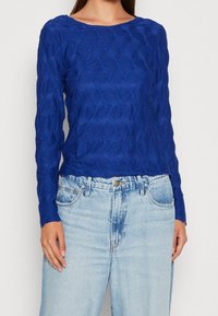 Blå strukturerad långärmad topp med ett mönster som liknar diamanter, i kombination med högmidjade ljusblå denimjeans.