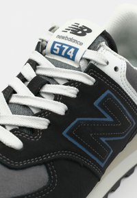 Black and gray sneaker with white laces, featuring a cushioned sole, mesh panels, and a blue accent on the side. Model 574 by New Balance.
