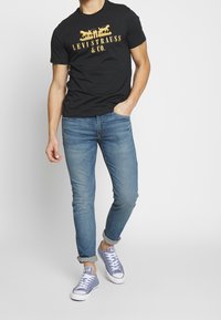 Camiseta negra de manga corta con el logo dorado "Levi Strauss & Co.", combinada con jeans ajustados de color azul y zapatillas de deporte azul claro.
