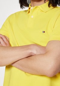 Keltainen polo-paita tekstuurikankaasta, jossa on kolme nappia; rintaan on brodeerattu pieni monivärinen logo.