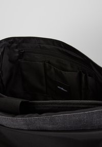 Interno di una borsa nera con una tasca con zip e numerosi scomparti. Il tessuto è liscio con una leggera trama. Etichetta del marchio visibile.