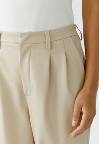 Beige plissierte Lederhosen mit glatter Textur, flachem Bund und seitlichen Taschen, neben einer Hand präsentiert.