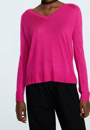 Persoon draagt een felroze, lange mouwen, V-hals trui en losse zwarte broek, met een zwarte choker ketting zichtbaar.