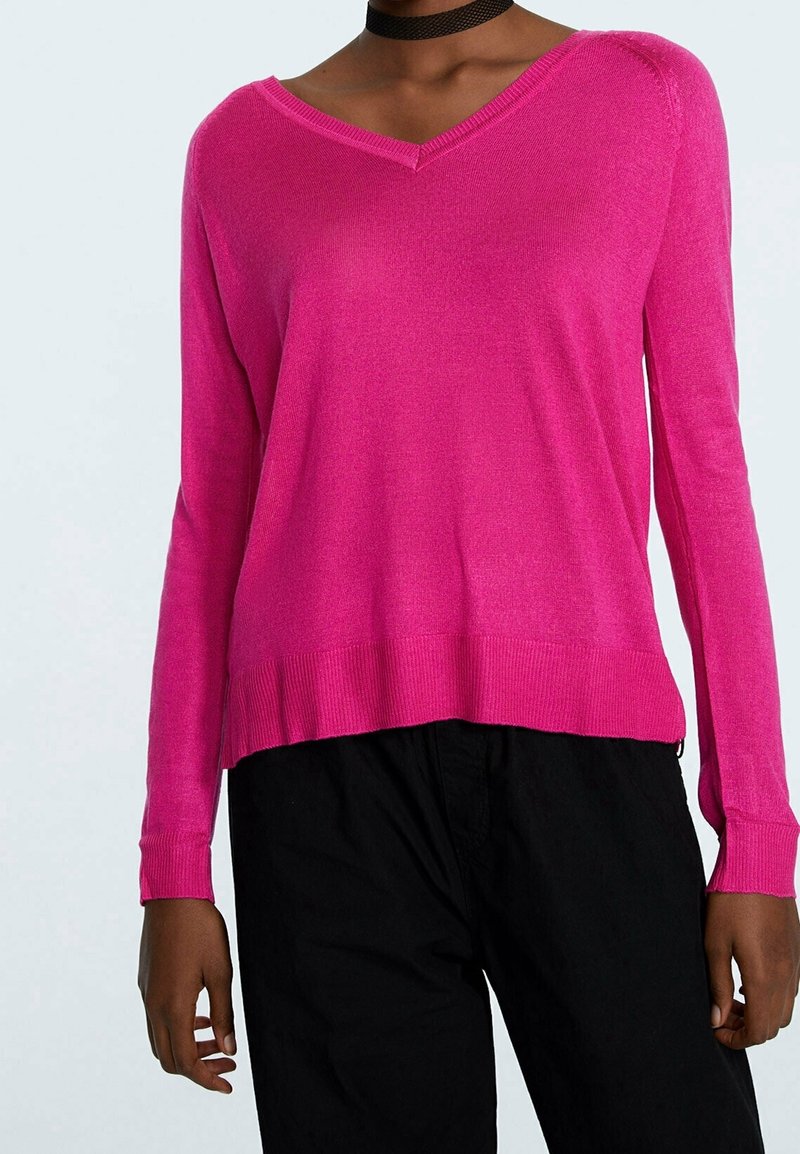 Personne portant un pull à manches longues rose vif avec un col en V et un pantalon noir ample, avec un collier ras du cou noir visible.