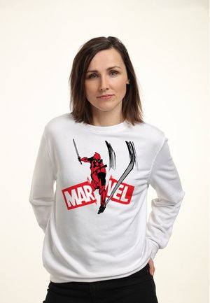 Kobieta o brązowych włosach ubrana w białą bluzę z nadrukiem Deadpoola i słowem Marvel w czerwieni i bieli na jednolitym jasnym tle.
