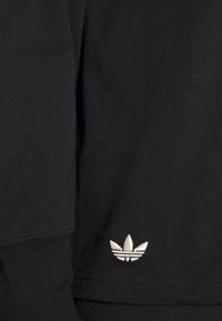 Černé bavlněné tričko s kulatým výstřihem a žebrovanými manžetami; na boku má malé vyšívané bílé logo ve tvaru tří listů.