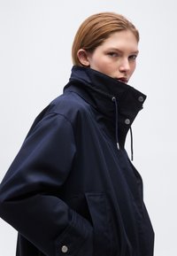 Γυναίκα με ανοιχτό καστανά μαλλιά φοράει μπλε σκούρο μπουφάν με ψηλό γιακά και πλαϊνές τσέπες μπροστά σε απλό φωτεινό φόντο.