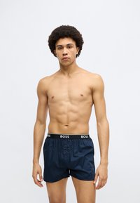 Marineblå bokser shorts med en svart elastisk linning som har merkevarenavnet "BOSS" i hvitt. Stoffet virker glatt, med én enkelt knappelukking.
