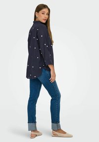 Navy oversized shirt with white heart patterns, long sleeves, and a high-low hem, paired with blue skinny jeans and beige slip-on shoes.