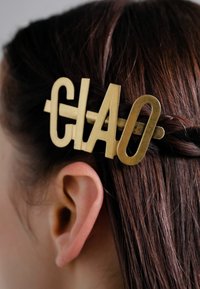 Brass hair clip with "CIAO" lettering; sleek design, matte finish, positioned in dark brown hair near the ear.