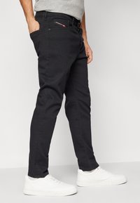 Homme portant un jean noir et des baskets blanches, se tenant avec une main dans la poche contre un fond clair uni.