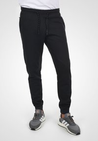 Sorte sweatpants med elastisk talje, snøre lukning og tapered fit. Parret med grå atletiske sko med hvide detaljer.