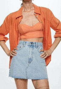 Camisa de lino naranja con botones sobre un top sin mangas durazno, combinada con una mini falda de mezclilla clara con dobladillo deshilachado y costura clásica.