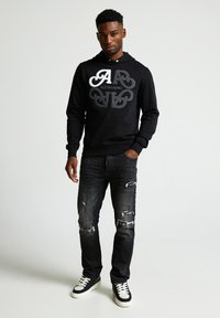 Sudadera negra con un logo plateado, combinada con jeans oscuros desgastados y zapatillas negras con suelas blancas. Diseño simple, corte casual.